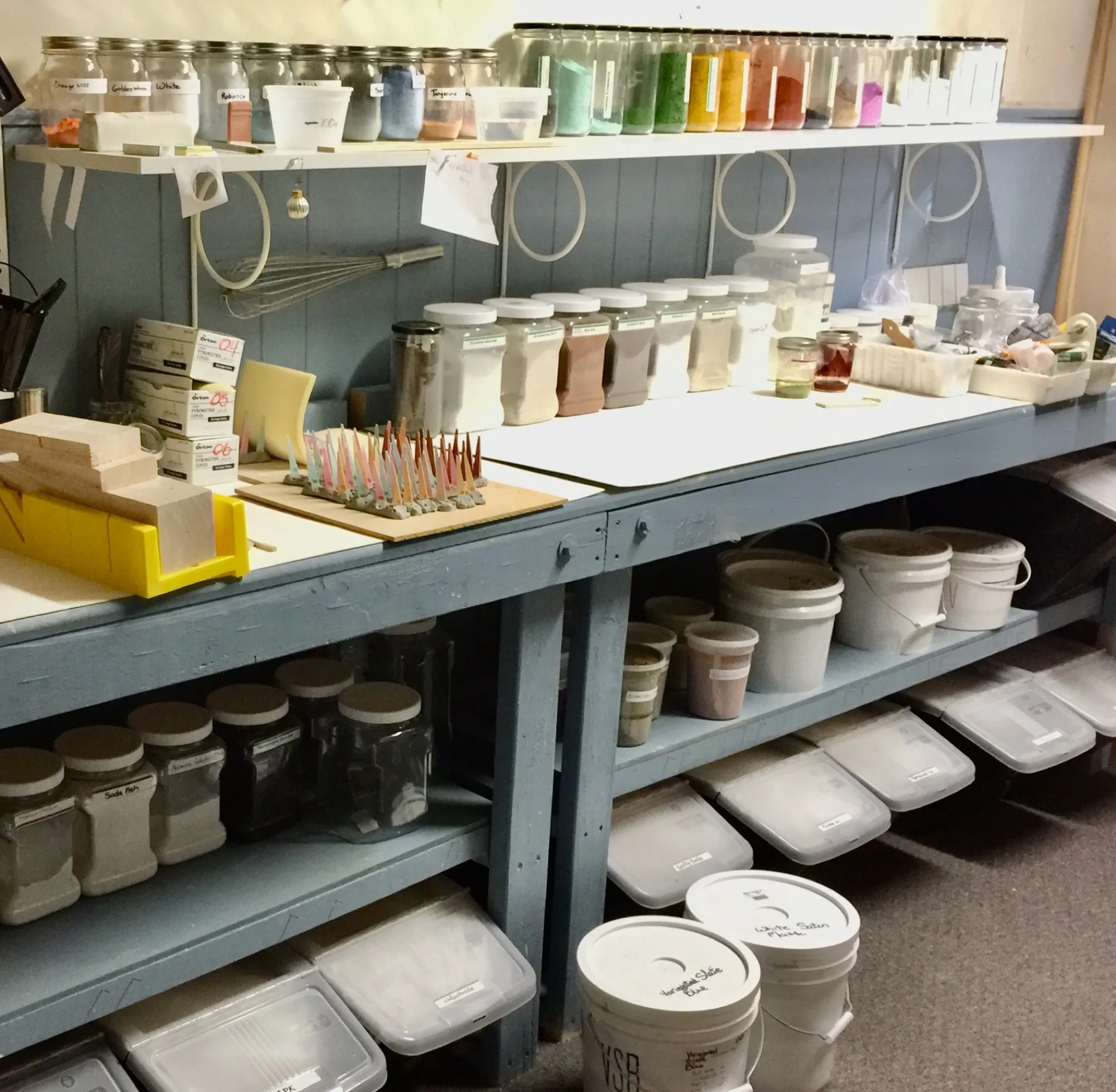 Shelves of labeled glaze materials and colorants in jars and buckets